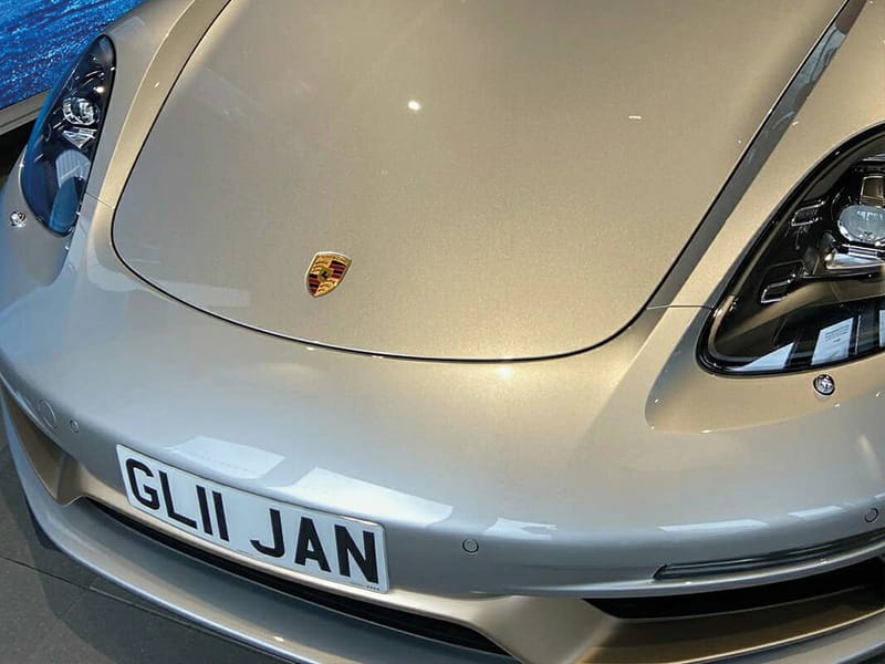 Close-up front view of a silver Porsche 718 Boxster, highlighting the bonnet, Porsche crest, headlight detail and compact white number plate displaying “GL11 JAN.”