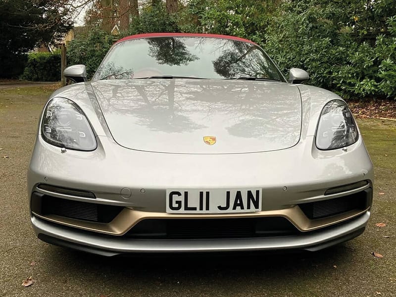 Front view of a silver Porsche 718 Boxster, showing the Porsche crest, large front air intakes and a compact white front number plate displaying “GL11 JAN.”