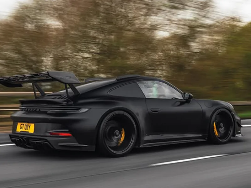 Side-rear rolling shot of a black Porsche 911 GT3, featuring a large rear wing, yellow brake calipers and a yellow shaped number plate displaying “GT 01YY.”