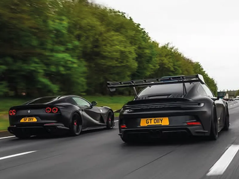 Rear rolling shot of a black Porsche 911 GT3 driving alongside a Ferrari, featuring a large rear wing and a yellow shaped number plate displaying “GT 01YY.”