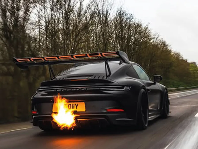 Rear three-quarter action shot of a black Porsche 911 GT3 with a large rear wing, showing flames from the exhaust and a yellow shaped number plate displaying “GT 01YY.”