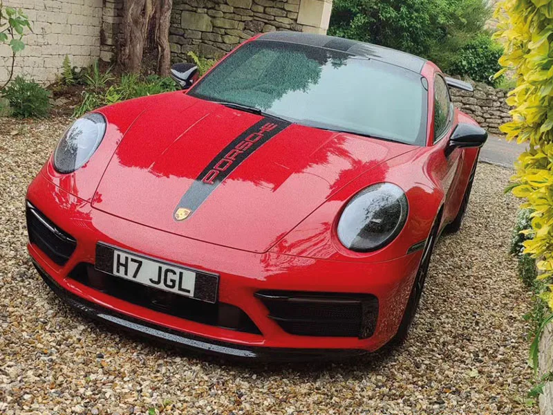 Front three-quarter view of a red Porsche 911, featuring a black Porsche bonnet stripe, dark headlight surrounds and a white front number plate displaying “H7 JGL.”