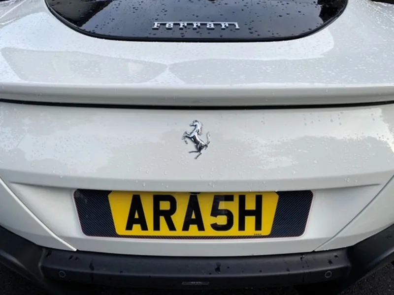Close-up rear view of a white Ferrari Roma, featuring Ferrari badging and a yellow carbon fibre-effect shaped number plate displaying “ARA5H.”