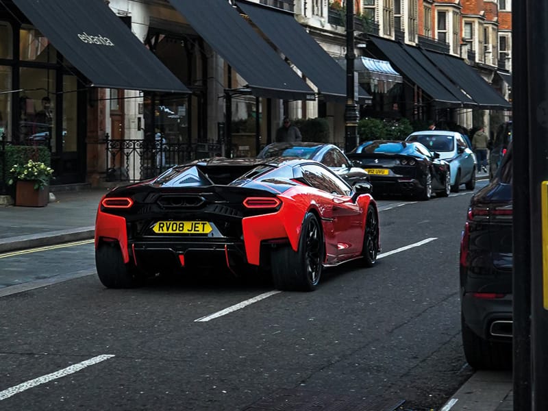 Rear view of a red Lamborghini Temerario driving through a city street, featuring distinctive LED taillights, rear diffuser and yellow shaped number plate displaying “RV08 JET.”