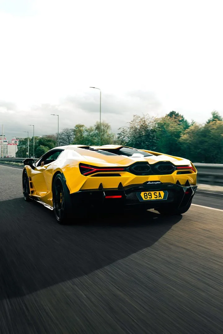 Rear view of a yellow Lamborghini sports car driving on a highway, with motion blur on the road and trees, under a cloudy sky. The car has black aerodynamic accents, distinctive angular taillights, and a visible rear engine cover.