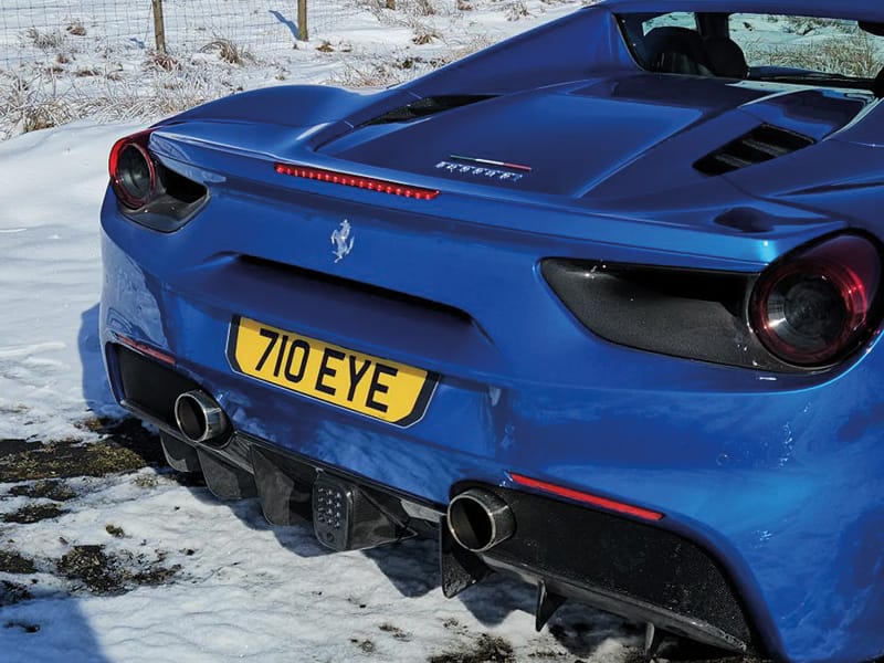 Rear three-quarter view of a blue Ferrari 488 in the snow, highlighting the sculpted rear bodywork, twin exhausts and yellow “710 EYE” number plate.