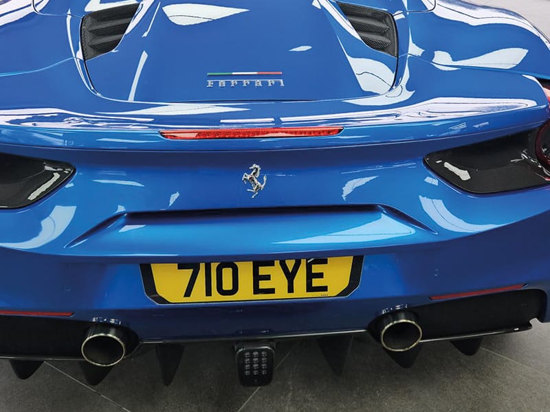 Close-up rear view of a blue Ferrari 488, showing the Ferrari badge, twin exhausts, rear diffuser and a yellow shaped number plate displaying “710 EYE.”