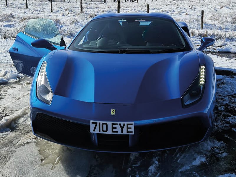 Front view of a blue Ferrari 488 parked in snowy conditions, featuring LED headlights and a compact white number plate displaying “710 EYE.”