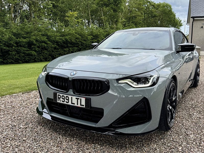 Front three-quarter view of a grey BMW M2, showing black kidney grilles, LED headlights, sporty front bumper detailing and a white number plate displaying “R99 LTL.”