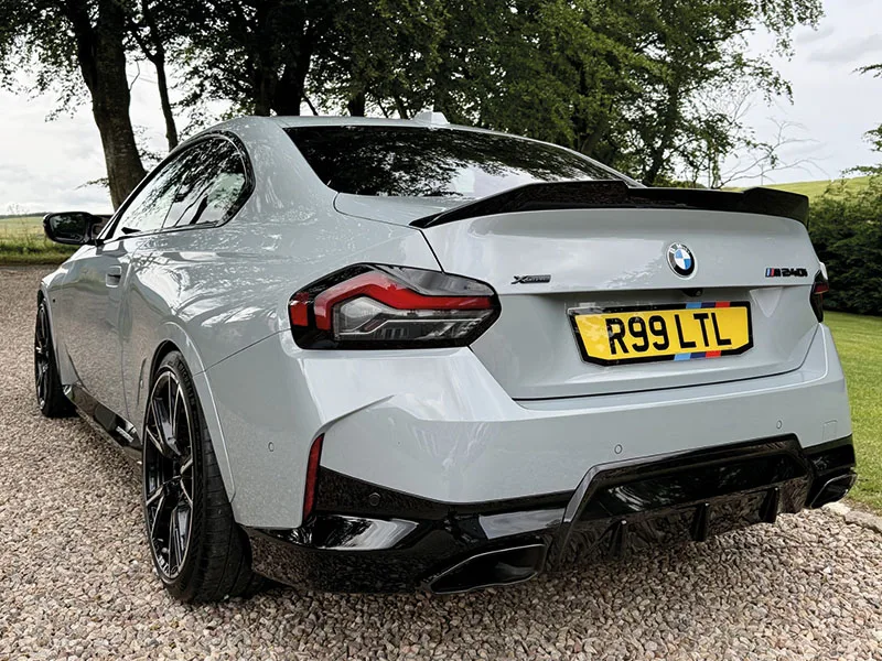 Rear three-quarter view of a grey BMW M2 fitted with a rear spoiler, black diffuser and yellow M Sport-themed number plate displaying “R99 LTL.”