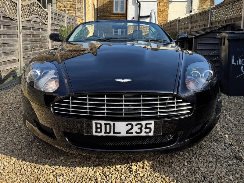 Front view of a black Aston Martin DB9 Volante parked on a gravel driveway, showing its chrome grille, rounded headlights and white front number plate “BDL 235.”