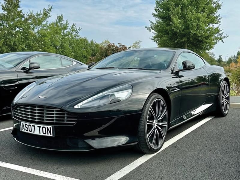 Front three-quarter view of a black Aston Martin DB9 parked in a car park, featuring its distinctive grille, swept headlights, multi-spoke alloy wheels and white UK registration plate “AS07 TON.”