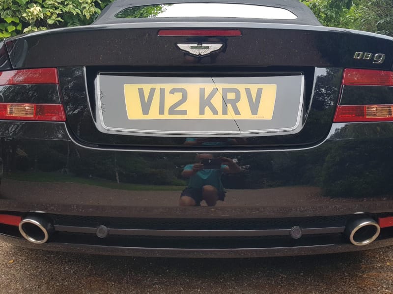 Close-up rear view of a black Aston Martin DB9 Volante, showing red taillights, twin exhausts and a yellow number plate displaying “V12 KRV.”