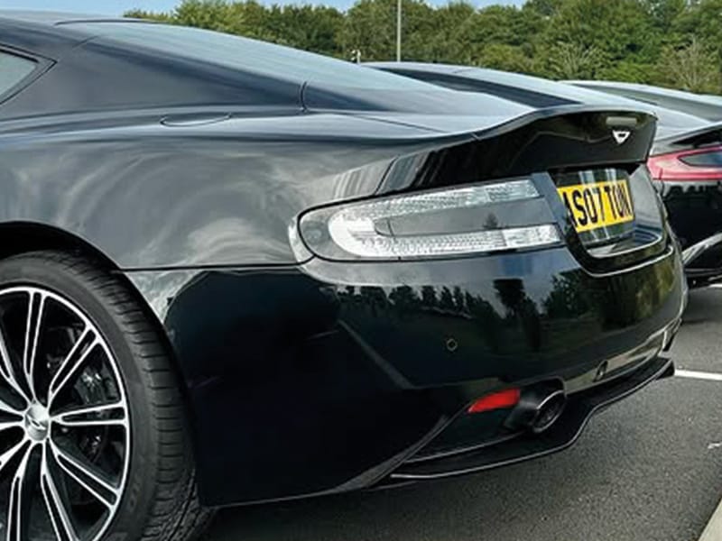 Close-up rear three-quarter view of a black Aston Martin DB9, highlighting the rear spoiler, clear taillight design, rear bumper and yellow “AS07 TON” number plate.