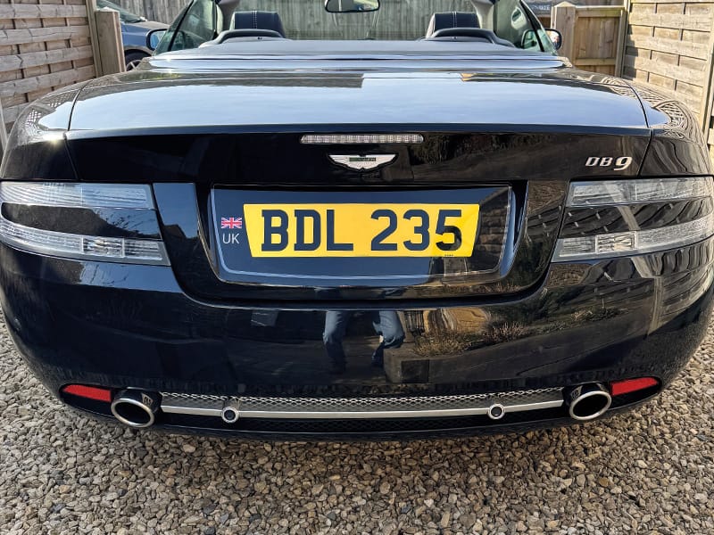 Rear view of a black Aston Martin DB9 Volante, featuring clear taillights, twin exhausts and a yellow UK number plate with Union Jack identifier displaying “BDL 235.”