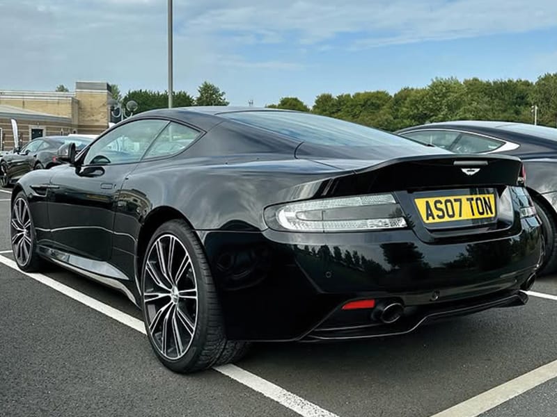 Rear three-quarter view of a black Aston Martin DB9, showing the sculpted rear spoiler, clear taillights, alloy wheels and yellow UK registration plate “AS07 TON.”