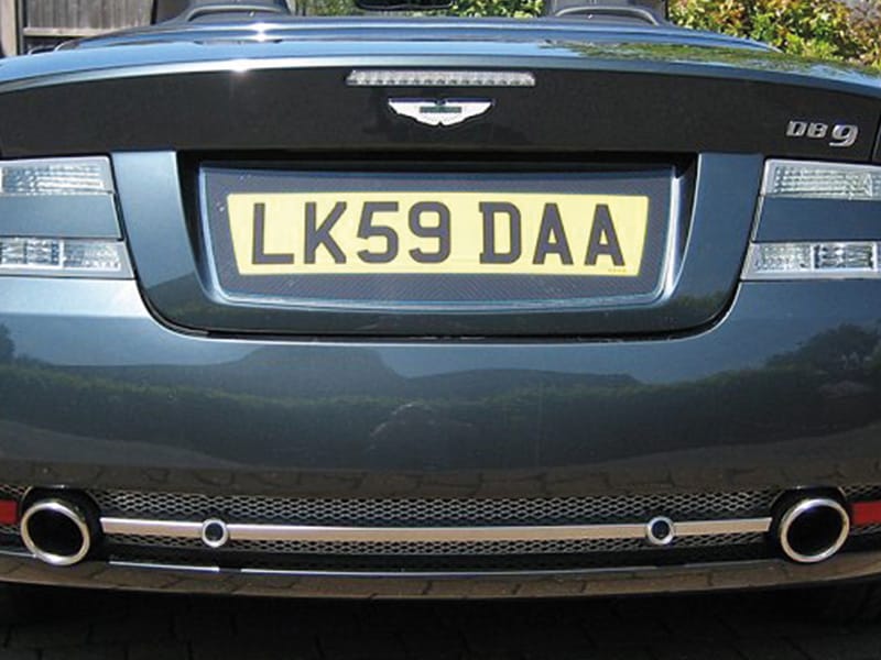 Close-up rear view of a dark Aston Martin DB9, featuring clear taillights, twin exhausts and a yellow carbon fibre-effect number plate displaying “LK59 DAA.”